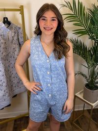 Woman wearing a blue floral romper standing in a room with a plant and clothing rack.