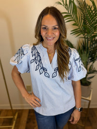 Woman wearing a white blouse with blue embroidery in a room with a plant