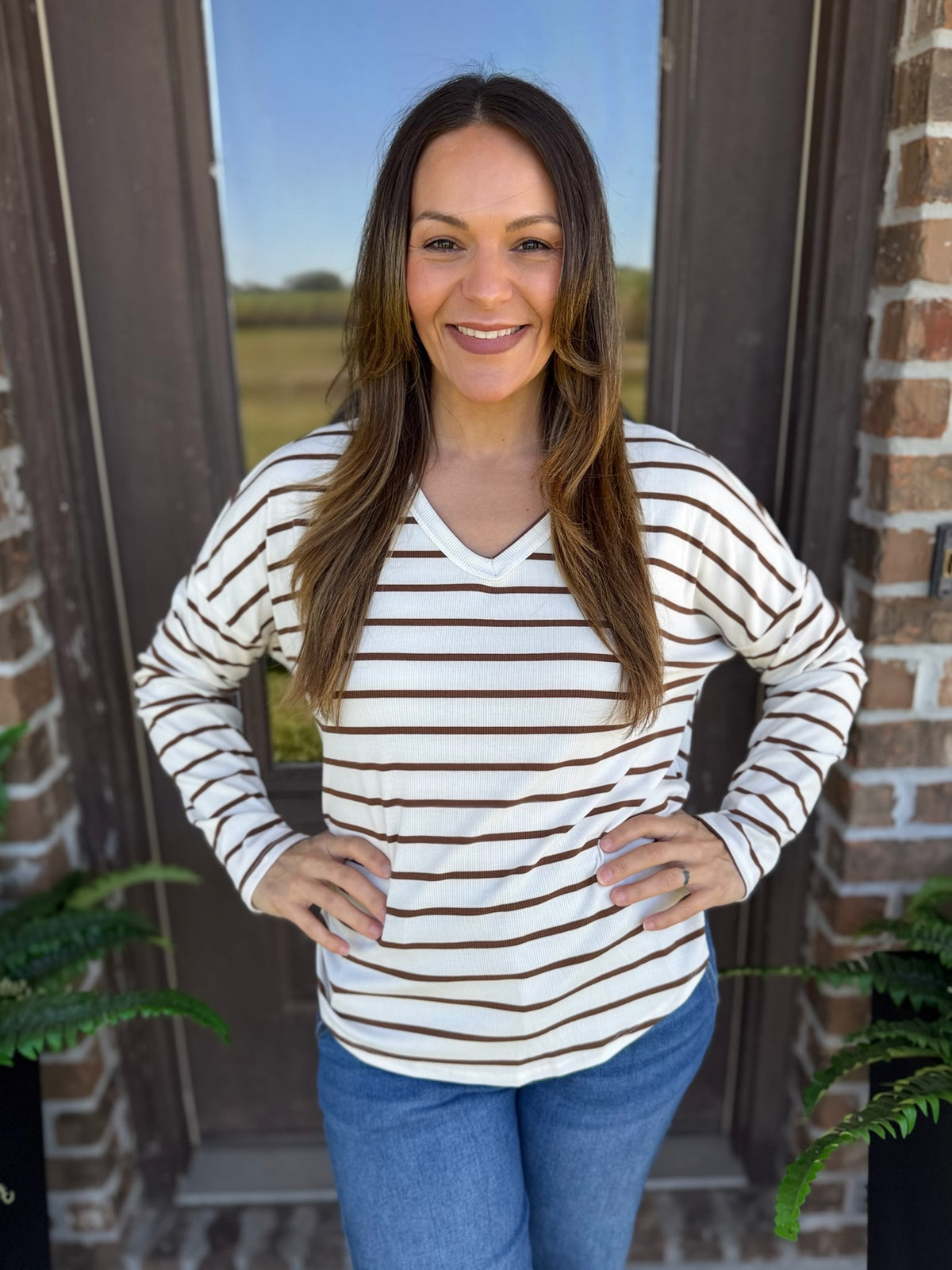 A woman with long brown hair stands in front of a brick door wearing a long-sleeve V-neck top with thin brown horizontal stripes on an off-white background, paired with blue jeans.