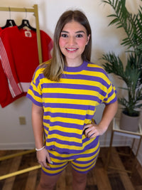 A woman modeling a matching two-piece lounge set consisting of a short-sleeve t-shirt and shorts, both featuring horizontal yellow and purple stripes. She is standing in a room with a wooden floor, a potted plant, and a clothing rack with a red shirt in the background.