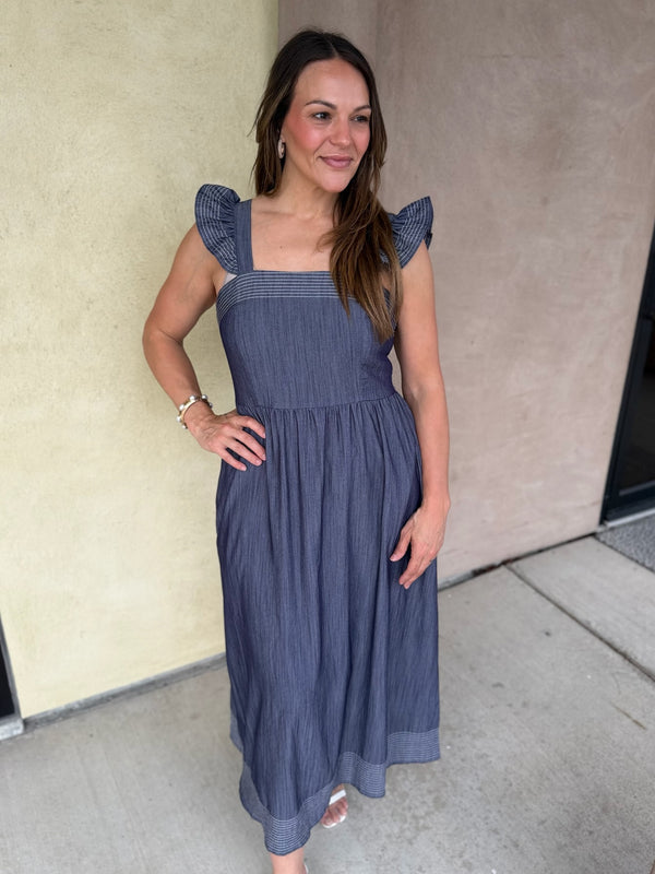 A woman wearing a slate blue pinstripe trimmed midi dress with ruffled cap sleeves, standing outdoors against a beige stucco wall.
