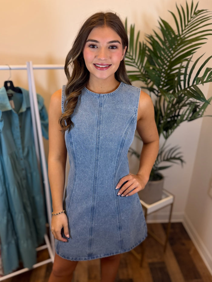 Woman wearing structured light wash denim mini dress with seam details in a room with plant and clothing rack in background.