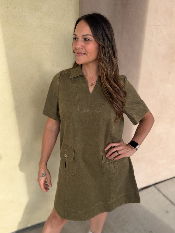 A woman modeling an olive green short-sleeve collared dress with front patch pockets, standing against a beige stucco wall.