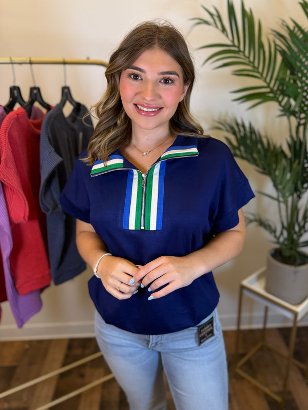 A woman wearing a navy blue short-sleeve top with a green, white, and blue striped collar and zipper detail, paired with light blue jeans, standing in a boutique setting.