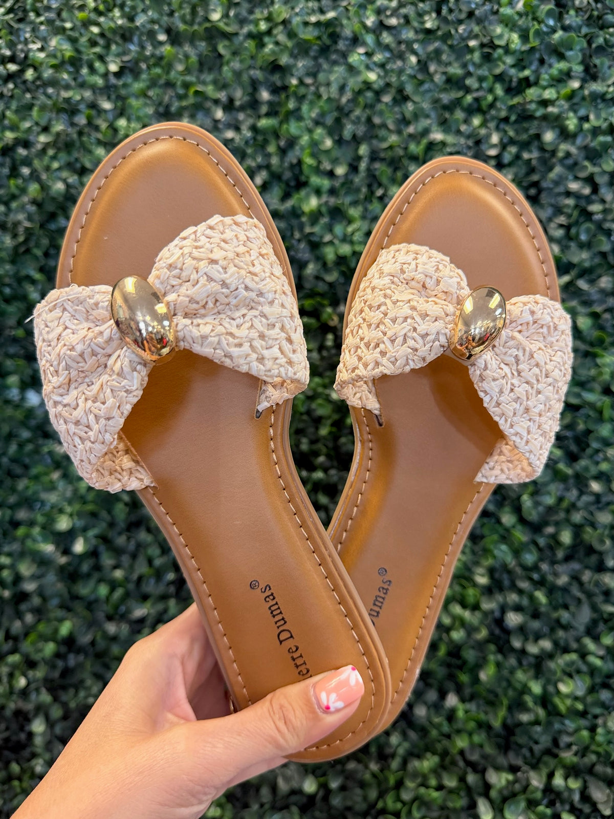 Pair of sandals with neutral textured bow details held against a green leafy background