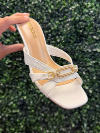 White sandal with gold chain detail held against a green leafy background