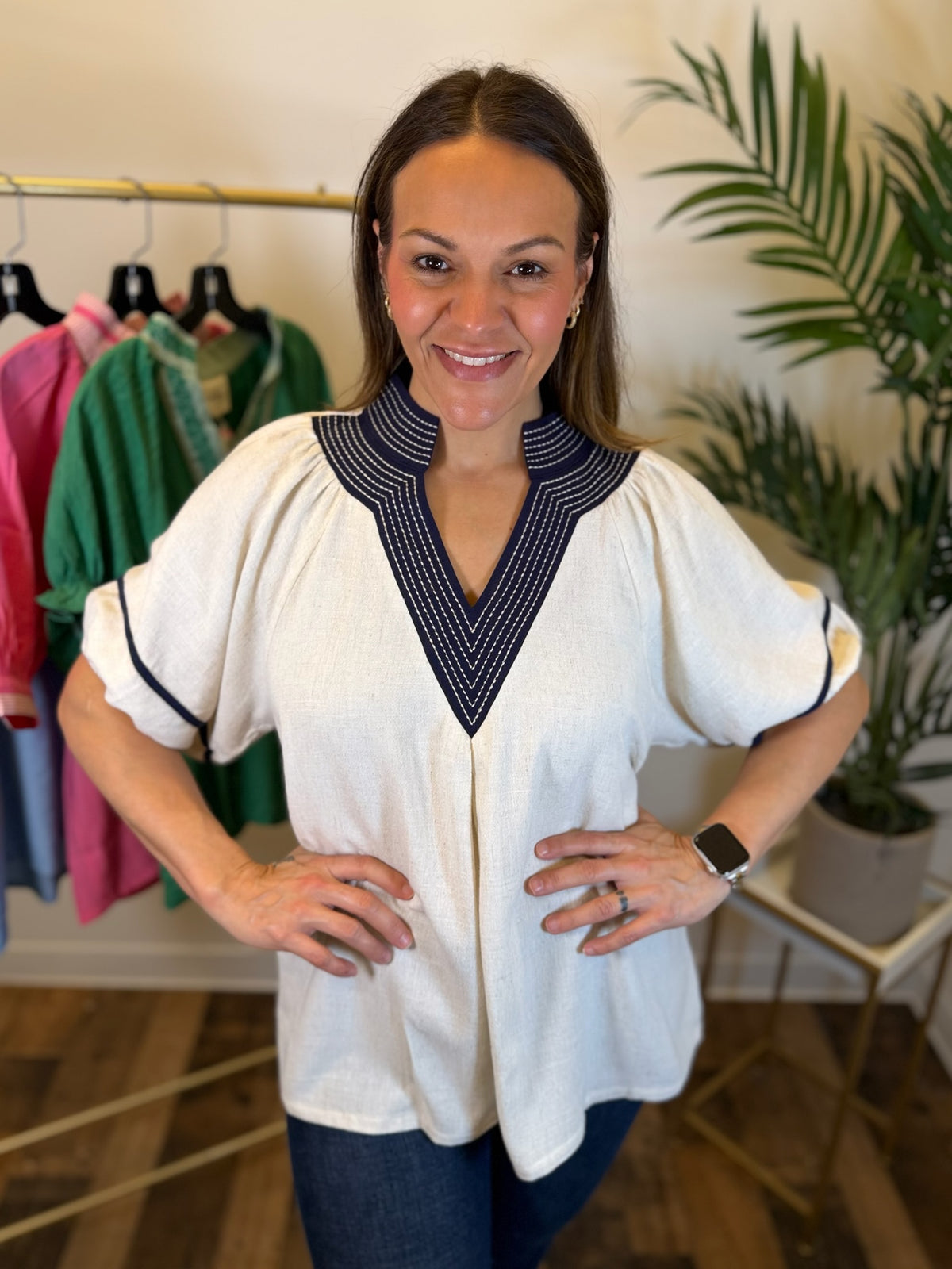 A woman modeling a short-sleeve cream blouse with a navy blue and white patterned V-neck collar and matching trim on the sleeve cuffs, paired with dark blue jeans in a boutique setting.