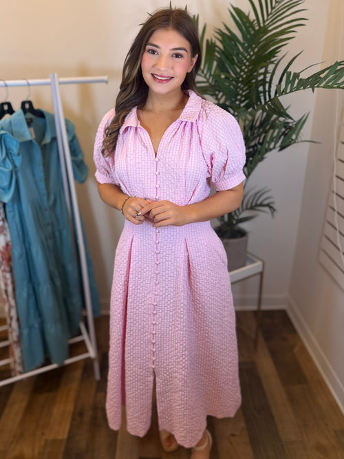 Woman wearing baby pink midi dress with textured fabric and puff sleeves in clothing store setting.