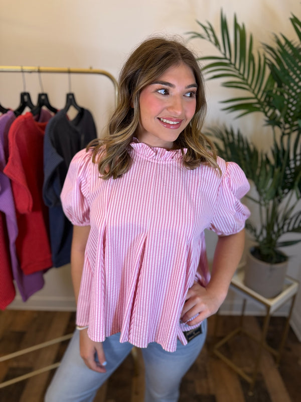 A woman wearing a pink vertically striped puff-sleeve blouse with a ruffled high neckline, paired with light blue jeans, standing in a boutique setting.