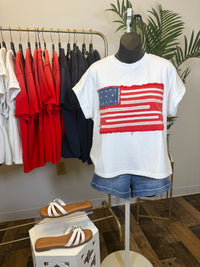 Mannequin wearing denim shorts and white short sleeve top with textured American flag applique hanging in a room with plant and clothing rack.