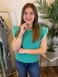 Woman wearing a teal knit top with white trimmed and ruffled sleeves in a room with plants and clothing racks.
