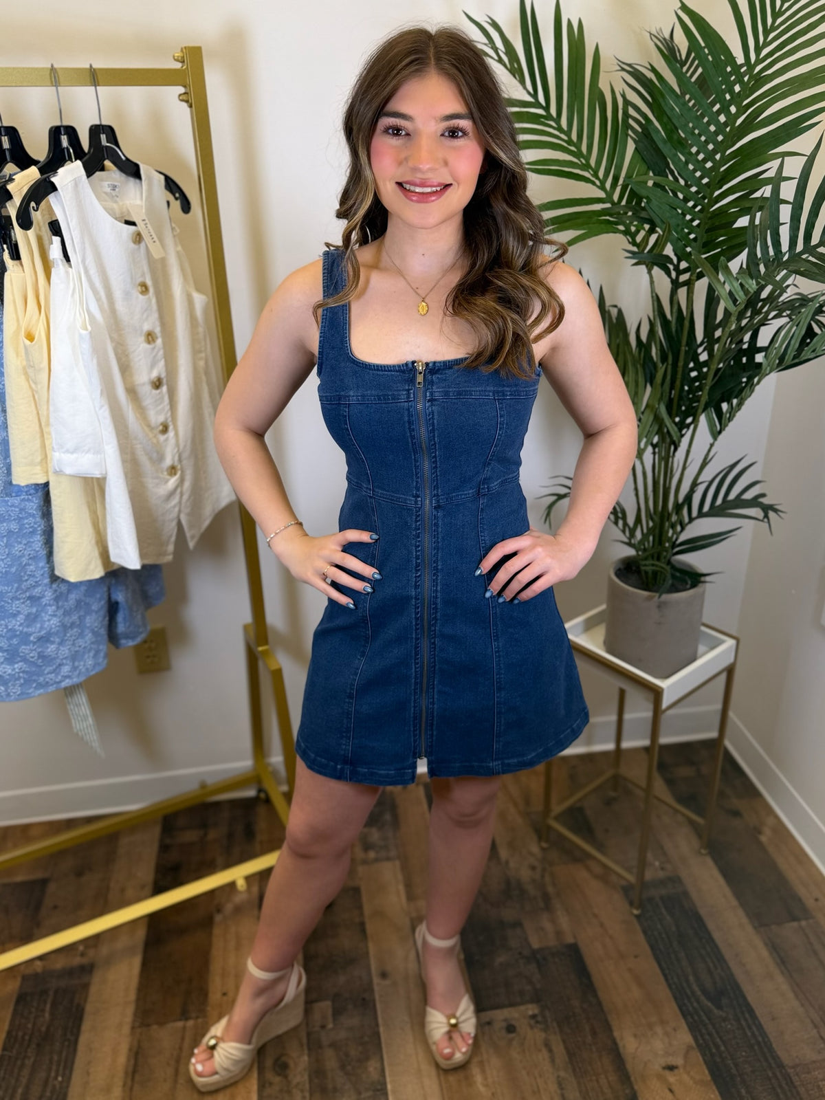 Woman wearing a blue denim dress standing in a room with a rack of clothes and a plant.