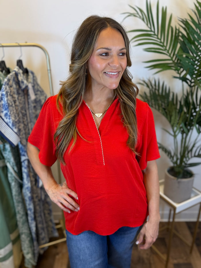 Woman wearing a red short sleeve blouse with scalloped collared neckline in a clothing store with plants in the background