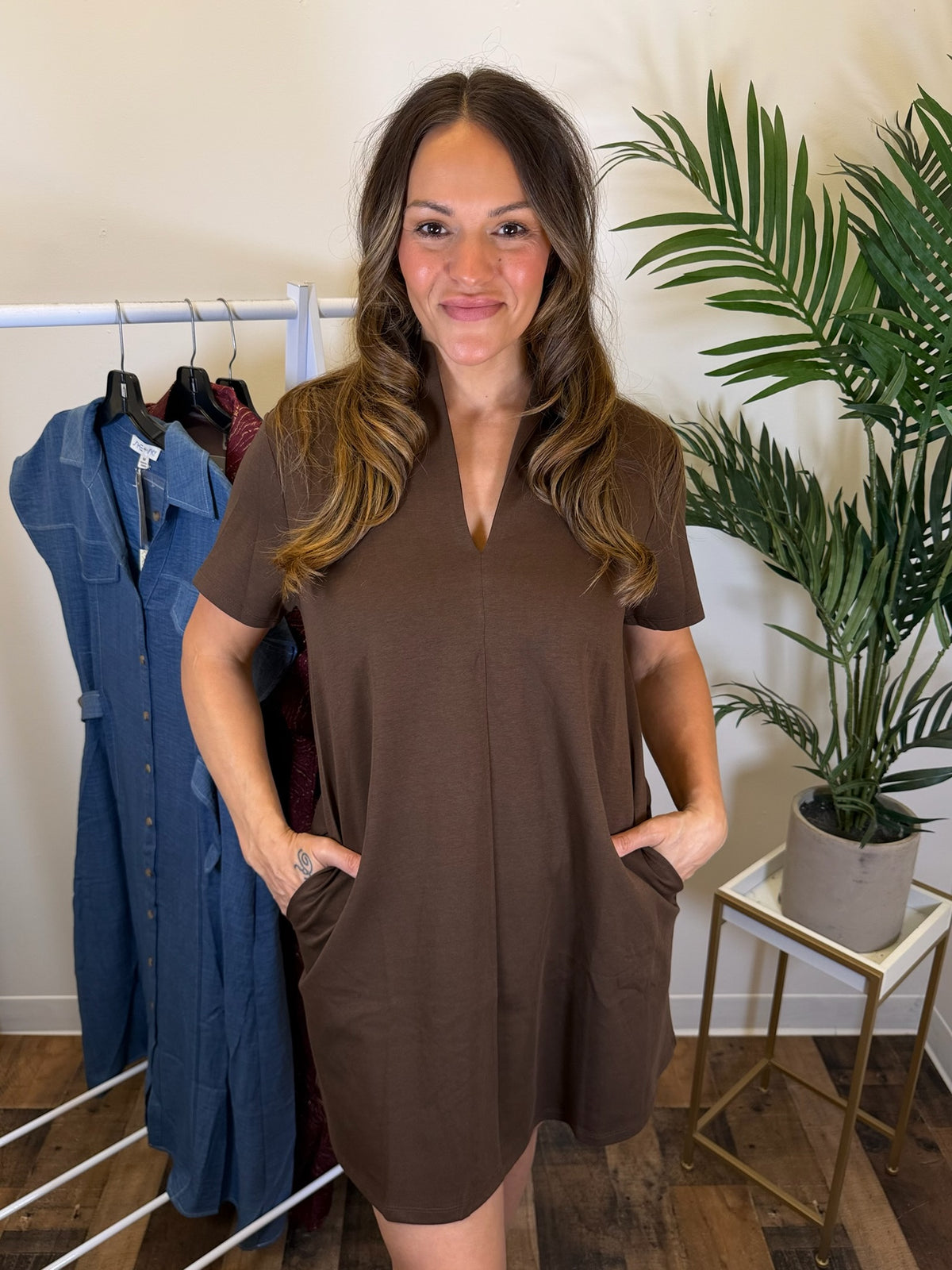A woman modeling a brown short-sleeve V-neck mini dress with side pockets, standing in a boutique setting next to a clothing rack and a potted plant.