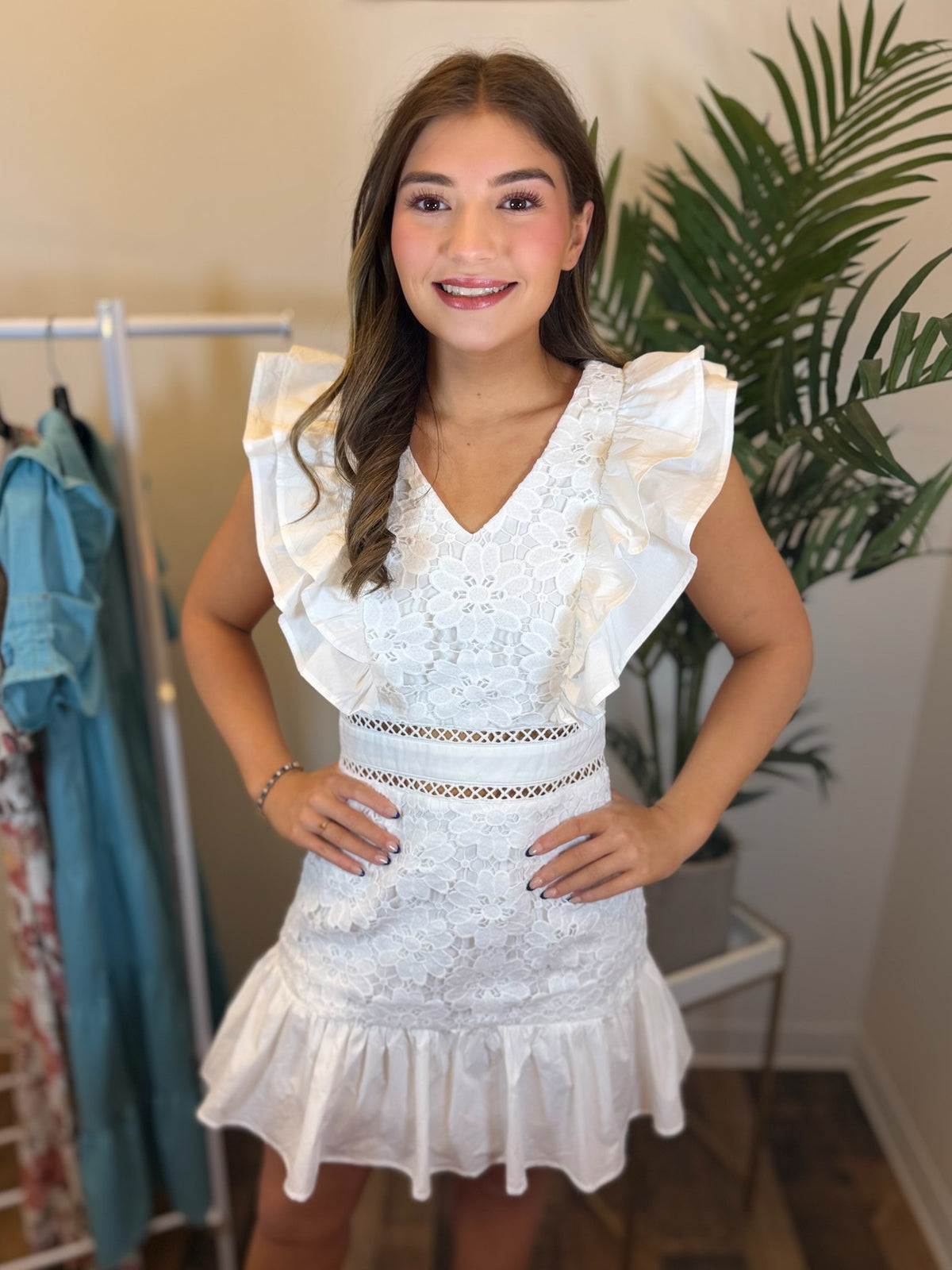Woman wearing a white lace dress with ruffled sleeves in a room with plants and clothing racks.