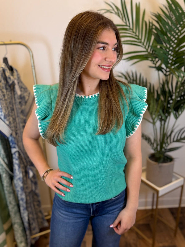 Woman wearing a teal knit top with white trim and ruffled sleeves in a room with plants and clothing.
