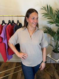 A woman wearing a light mocha short-sleeve collared shirt and dark blue jeans, standing in a boutique setting with a clothing rack and a potted plant in the background.
