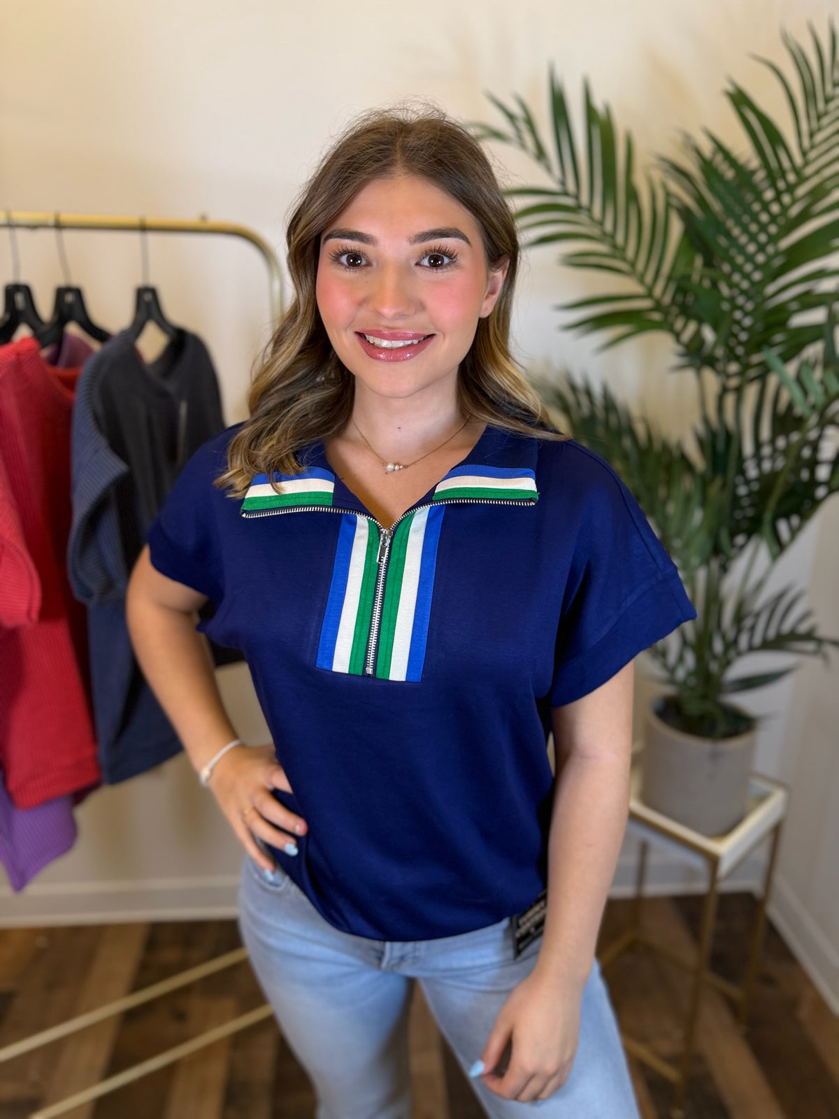 A woman wearing a royal blue short-sleeve top with a green, white, and blue striped collar detail, paired with light blue jeans, standing in a boutique setting.