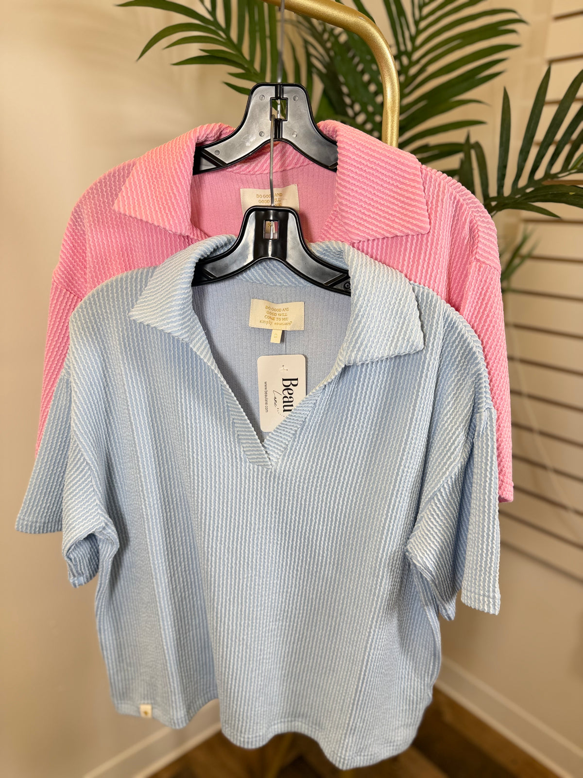 Two short sleeve ribbed shirts, one blue and one pink, hanging on a rack with a plant in the background.