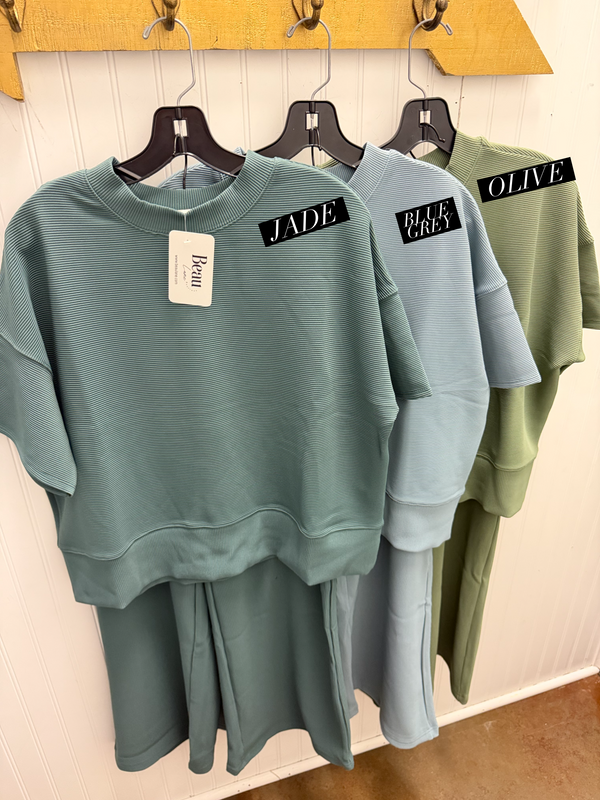 Three women's lounge sets in teal, light blue, and olive green hanging on black hangers against a white paneled wall. Each set includes a short-sleeve top and matching pants.