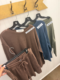 A set of ribbed knit lounge shorts and matching top in brown, hanging on a black hanger, with two other color options (blue and olive) hanging behind them on a wooden wall hook display. A hand is holding the brown pants in the foreground.