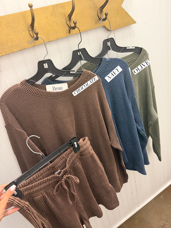 A set of ribbed knit lounge shorts and matching top in brown, hanging on a black hanger, with two other color options (blue and olive) hanging behind them on a wooden wall hook display. A hand is holding the brown pants in the foreground.