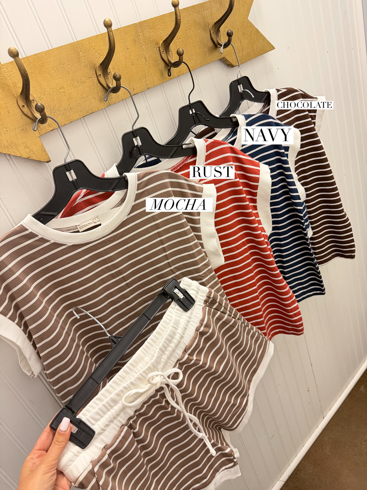 A set of women's two-piece lounge sets in various stripe patterns and colors, hanging on black hangers against a white paneled wall with a gold hook rack. The sets feature mocha and white stripes, rust and white stripes, navy and white stripes, and brown and white stripes.